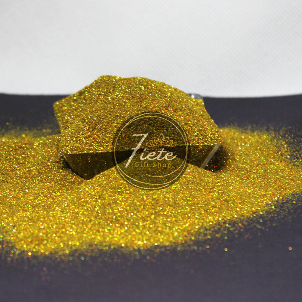 Extra fine glitter overflowing from a small clear container. The container is surrounded by the same glitters.  This glitter is holographic and gold, reflecting light showing all the colors of the rainbow. 